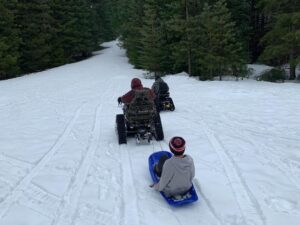 Two people in their own David's Chair on a snowy road pulling a kid in a sled
